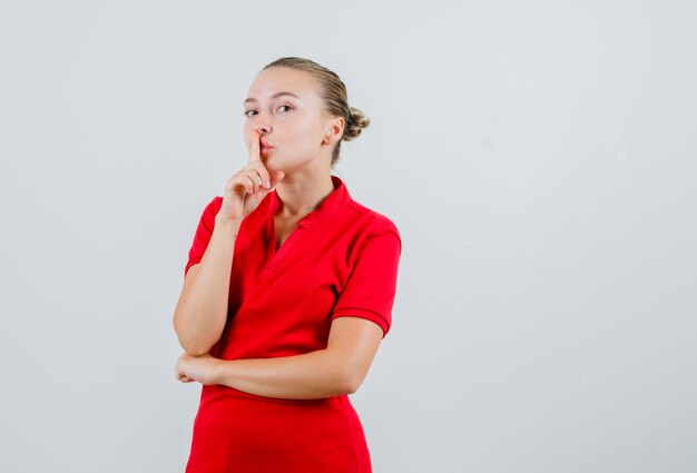 沈黙のジェスチャーを示し、注意深く見える赤いTシャツの若い女性