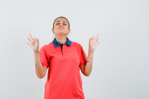 Young woman in red t-shirt standing in meditating pose and looking calm , front view.