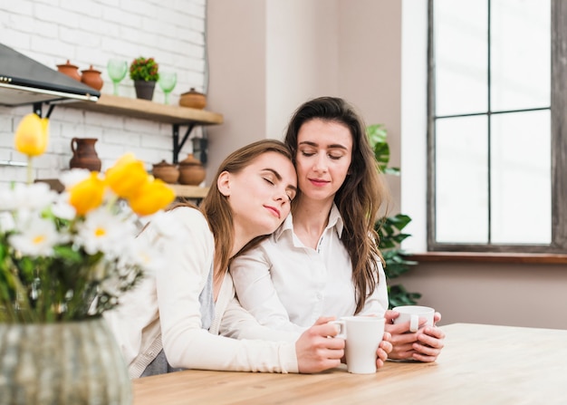 若い女性が手に一杯のコーヒーを保持している彼女のガールフレンドの肩でリラックス