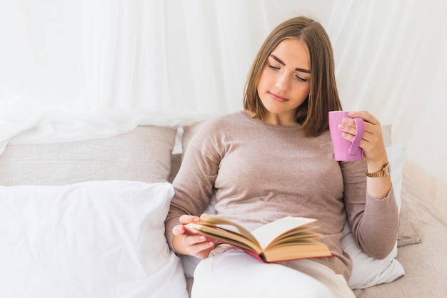 커피 컵을 손에 들고 책을 읽고 침대에서 편안한 젊은 여자