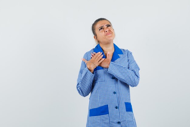 Young woman resting hands on chest, looking above in blue gingham pajama shirt and looking pensive. front view.