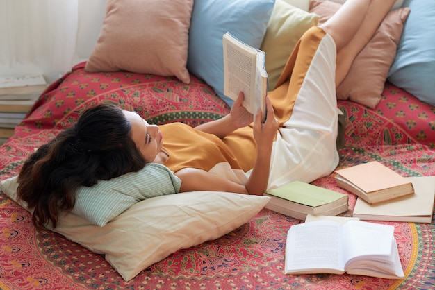 Young woman resting with book in bed with her legs on pillows