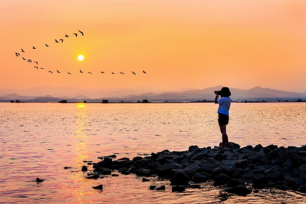 Young woman on the rocks waterfront taking photos of beautiful natural landscape birds flying in a row sun on the orange sky at sunset background by dslr camera, krasiao dam, suphan buri in thailand