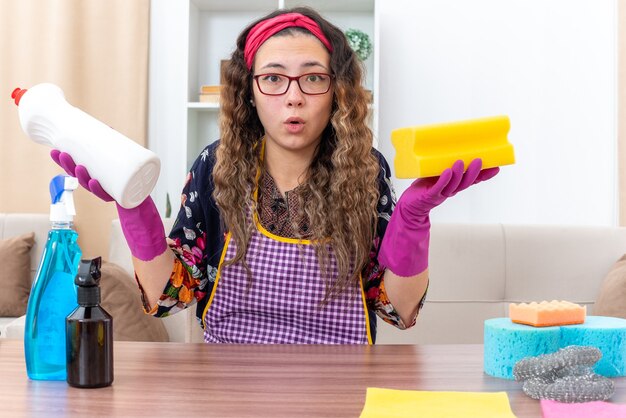 Young woman in rubber gloves holding sponge and bottle with cleaning supplies looking surprised sitting at the table with cleaning supplies and tools in light living room