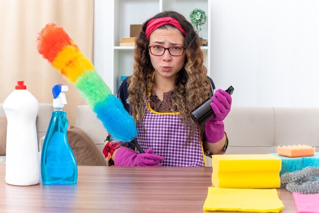 Young woman in rubber gloves holding static duster and cleaning spray looking confused and displeased sitting at the table with cleaning supplies and tools in light living room