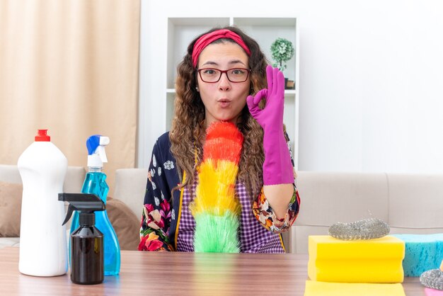 Young woman in rubber gloves holding static duster  happy and surprised doing ok sign sitting at the table with cleaning supplies and tools in light living room