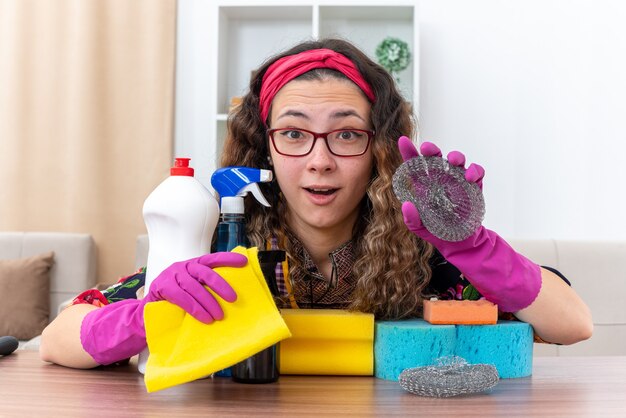 Young woman in rubber gloves looking at camera amazed and happy sitting at the table with cleaning supplies and tools in light living room