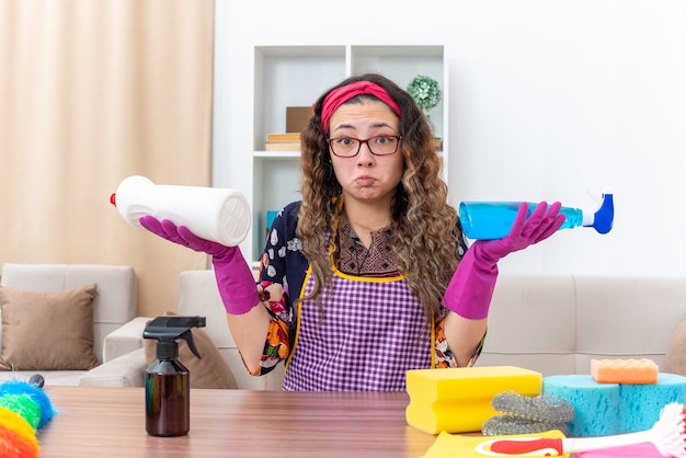 Foto gratuita giovane donna con guanti di gomma seduta al tavolo con prodotti per la pulizia e strumenti confusi cercando di fare una scelta in un soggiorno luminoso