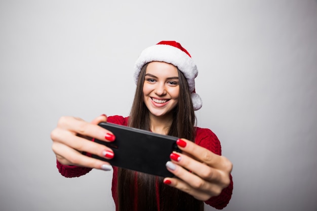 Giovane donna in cappello della santa prende selfie al telefono isolato sul muro grigio
