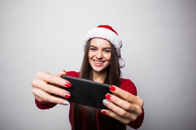 Young woman in santa hat take selfie on the phone isolated on gray wall