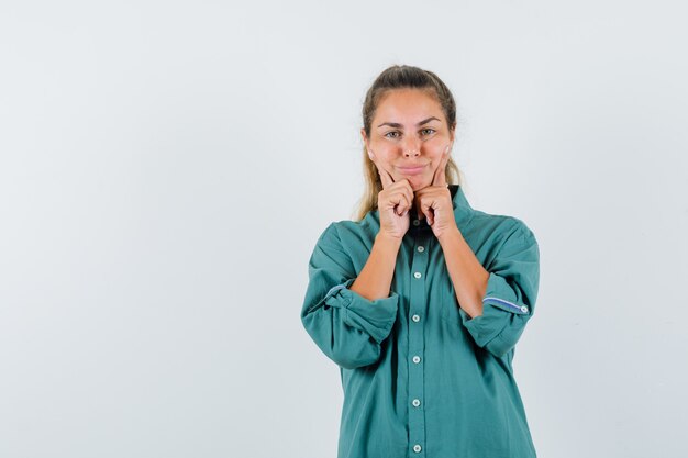 Young woman scratching head in green blouse in holding index fingers near mouth, forcing a smile and looking attractive