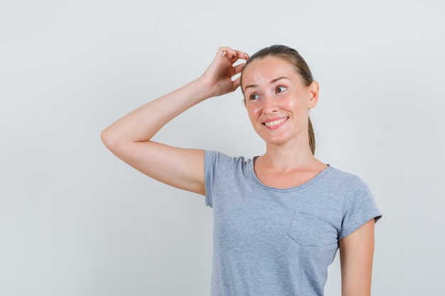 Young woman scratching head in grey t-shirt and looking hesitant. front view.