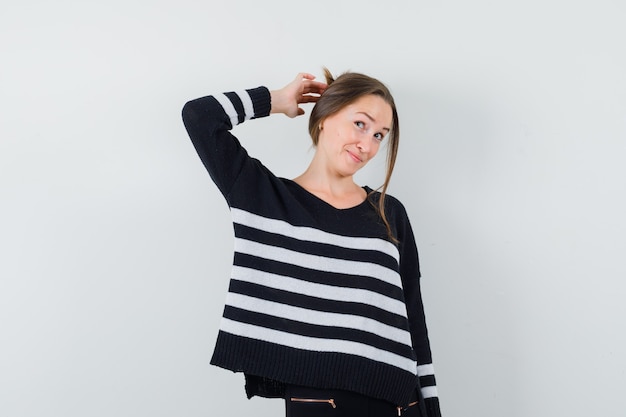 Young woman scratching head in striped knitwear and black pants and looking optimistic