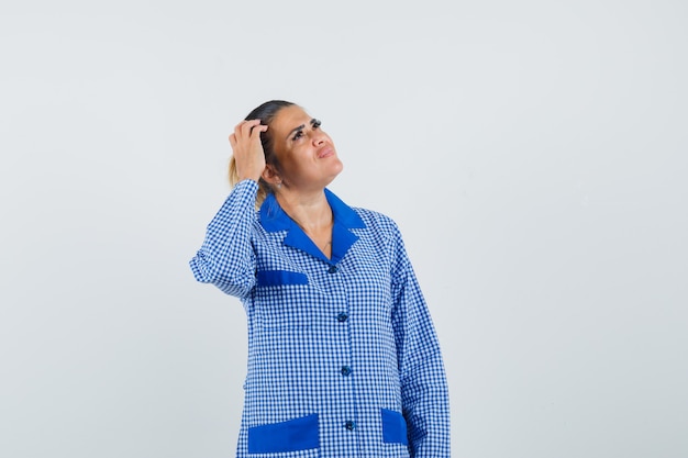 Free photo young woman scratching head while thinking about something in blue gingham pajama shirt and looking pensive. front view.