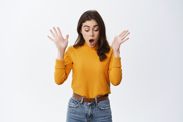 Young woman screams and looks down with shocked gasping face, drop something on floor and staring startled at bottom empty space, standing against white wall