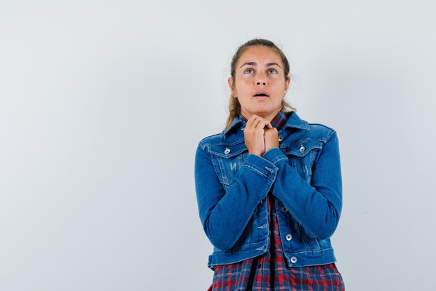 Young woman in shirt, jacket clasping hands in praying gesture and looking hopeful , front view.