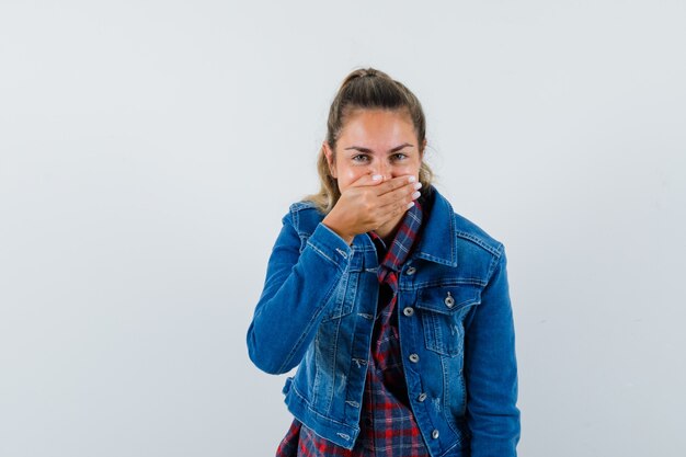 Young woman in shirt, jacket holding hand on mouth and looking glad , front view.