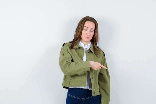 Young woman in shirt, jacket pointing down and looking optimistic, front view.