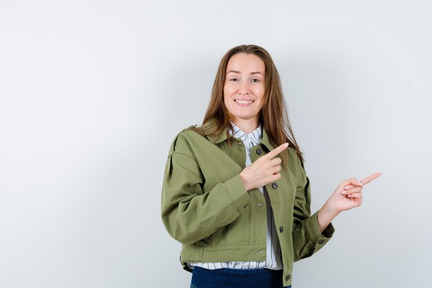 Young woman in shirt, jacket pointing at upper right corner and looking cheerful, front view.