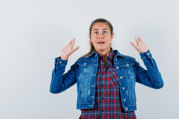 Young woman in shirt, jacket raising hands in surrender gesture and looking puzzled , front view.