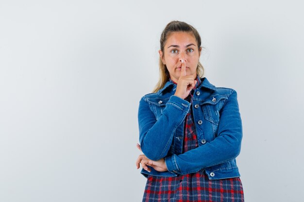 Young woman in shirt, jacket showing silence gesture and looking pensive , front view.