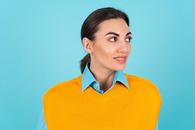 Молодая женщина в рубашке и оранжевом жилете на бирюзовом фоне задумчиво смотрит в сторону с легкой улыбкой на лице