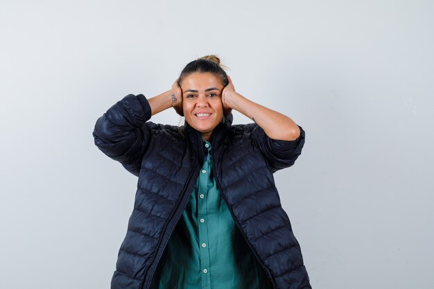 Young woman in shirt, puffer jacket clasping head with hands and looking glad , front view.