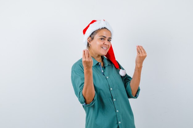 셔츠를 입은 젊은 여성, 산타 모자는 이탈리아 제스처를 보여주고 쾌활하고 앞모습을 보고 있습니다.