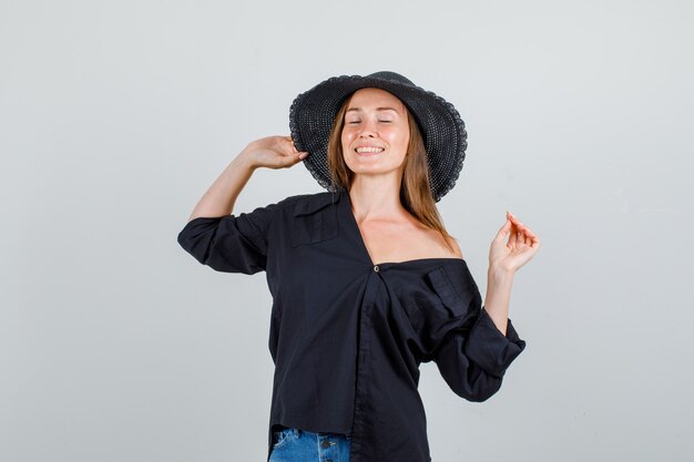 Young woman in shirt, shorts holding her hat with closed eyes and looking cheery