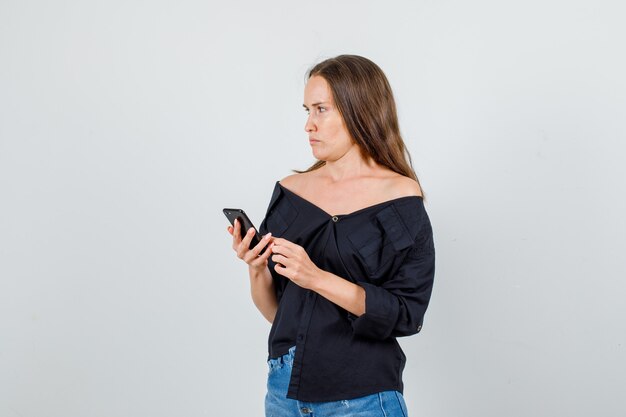 Young woman in shirt, shorts looking aside while holding smartphone and looking sad