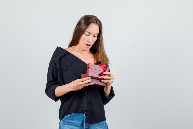 Молодая женщина в рубашке, шортах смотрит в подарочную коробку и выглядит удивленно