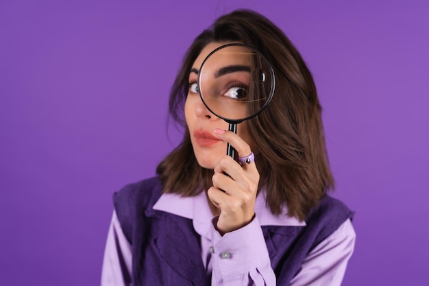 Молодая женщина в рубашке и жилете на фиолетовом фоне развлекается с увеличительным стеклом в руке
