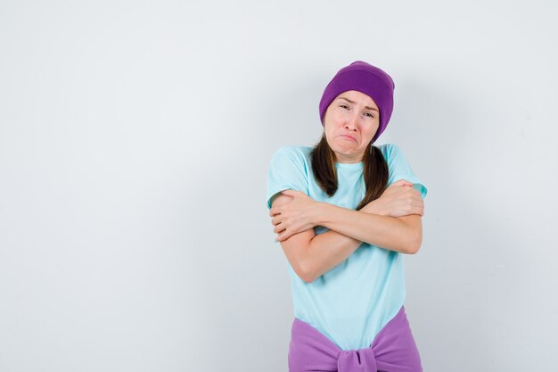 Young woman shivering from cold in blue t-shirt, purple beanie and looking exhausted. front view.