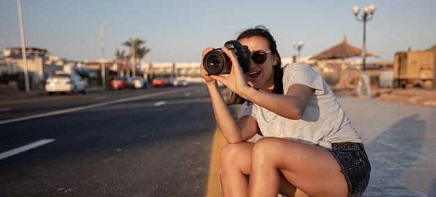 A young woman in shorts and a T-shirt in summer sits on the sidewalk with a professional camera