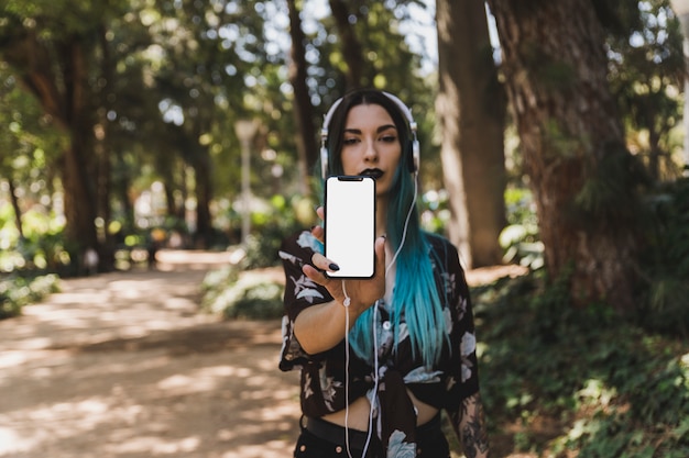 Молодая женщина показывает пустой белый смартфон с наушниками на голове