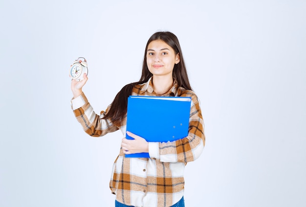 Young woman showing blue folder and clock on white wall. 