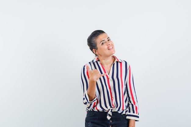 Young woman showing call me gesture in striped blouse and looking optimistic