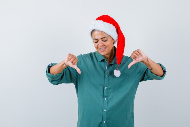 シャツ、サンタの帽子、陽気に見える、正面図で二重親指を示す若い女性。