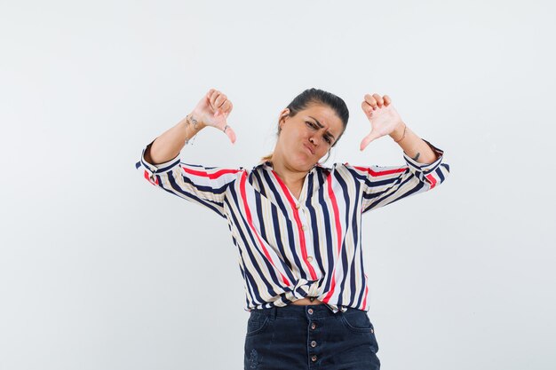 Young woman showing double thumbs down with both hands in striped blouse and looking displeased