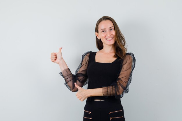 Young woman showing double thumbs up in black blouse and black pants and looking happy. front view.