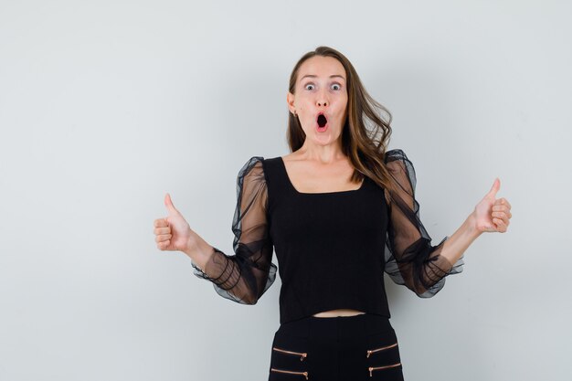 Young woman showing double thumbs up in black blouse and black pants and looking happy. front view.