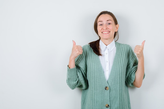 Young woman showing double thumbs up in blouse, cardigan and looking joyful , front view.