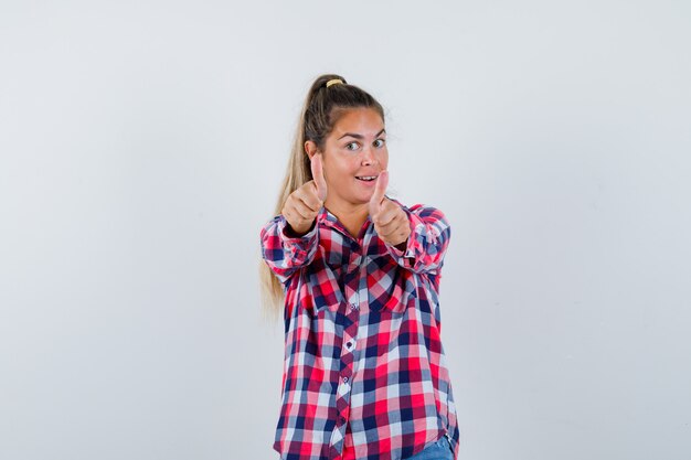 Молодая женщина показывает двойные пальцы вверх в повседневной рубашке и выглядит счастливой. передний план.