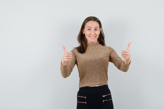 Young woman showing double thumbs up in gold gilded sweater and black pants and looking happy. front view.