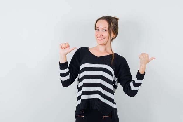 Young woman showing double thumbs up and pointing right in striped knitwear and black pants and looking happy