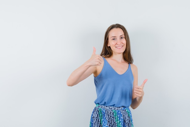 Young woman showing double thumbs up in singlet, skirt and looking joyful , front view.