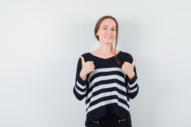 Young woman showing double thumbs up in striped knitwear and black pants and looking happy