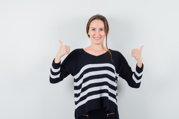 Young woman showing double thumbs up in striped knitwear and black pants and looking happy