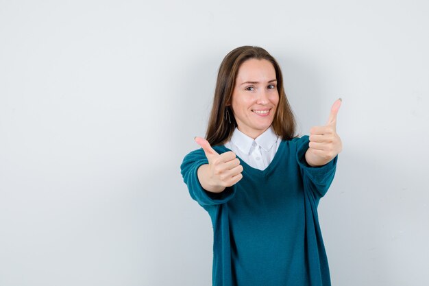 Молодая женщина показывает двойные пальцы вверх в свитере над белой рубашкой и выглядит счастливой, вид спереди.