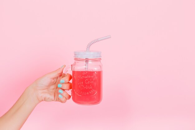 Young woman showing a glass with freshly squeezed berry juice, which she made for lunch with family. Photo of big cup with bright tasty lemonade for birthday party isolated on pink background